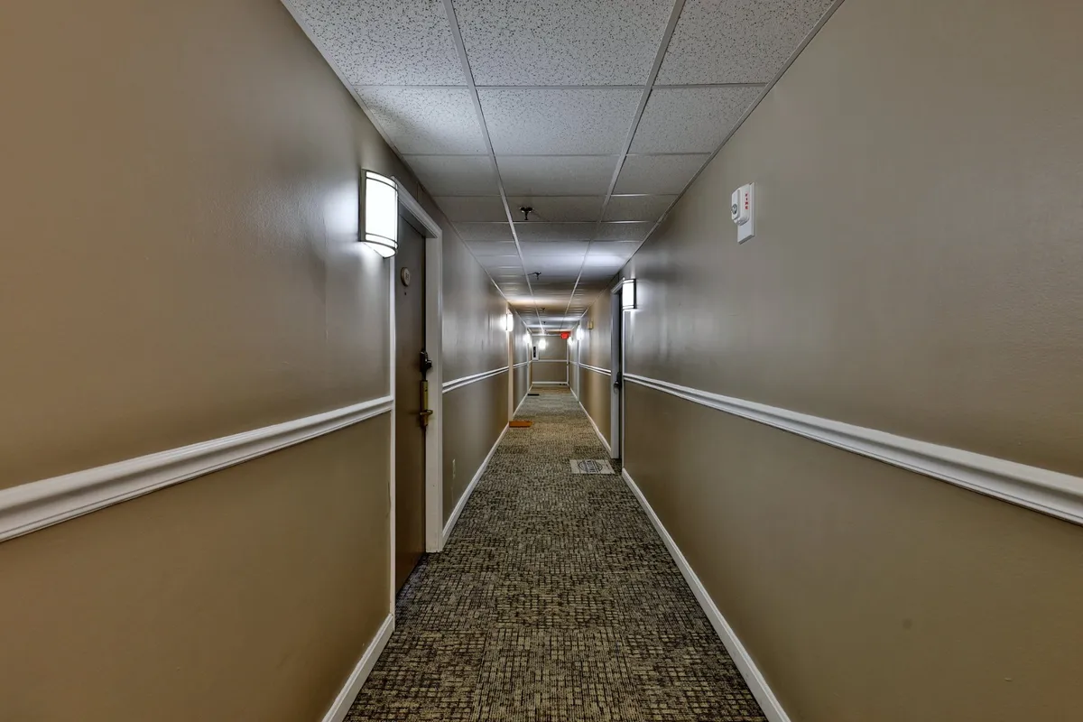 This image depicts an interior hallway with soft lighting, visible light fixtures, and several doors, giving a clear sense of building common areas and entryways. The carpeted texture and neutral decor suggest a welcoming and practical entry experience for guests.