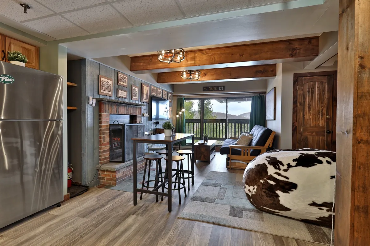 A dining area with a wooden table set near a fireplace, large windows revealing mountain views, and adjacent seating like a sofa and bean bag. The mood is cozy and inviting, perfect for group gatherings, meals, and conversations.