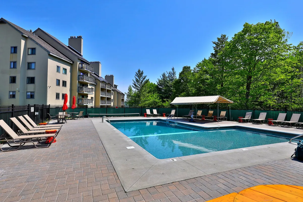 A vibrant exterior view of the outdoor heated swimming pool, surrounded by lounge chairs, lush greenery, and a stylish cabana area, evoking a resort-like, cheerful summer atmosphere.