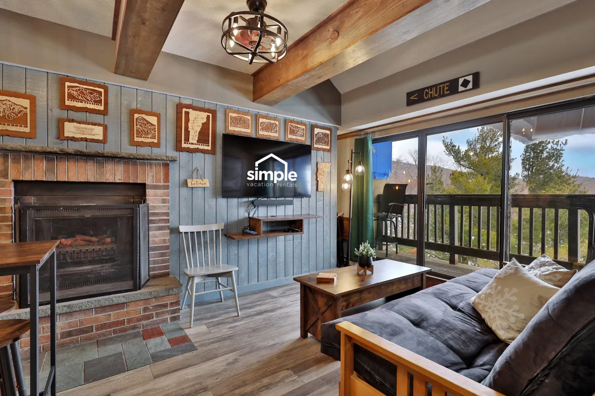 The hero image showcases a warm, inviting living space with a glowing fireplace as the centerpiece, complemented by comfortable wooden furnishings, tasteful wall art, and expansive windows that let in natural light and offer scenic views. The layout encourages relaxation, socializing, and enjoying both indoor comforts and glimpses of Vermont’s natural beauty.
