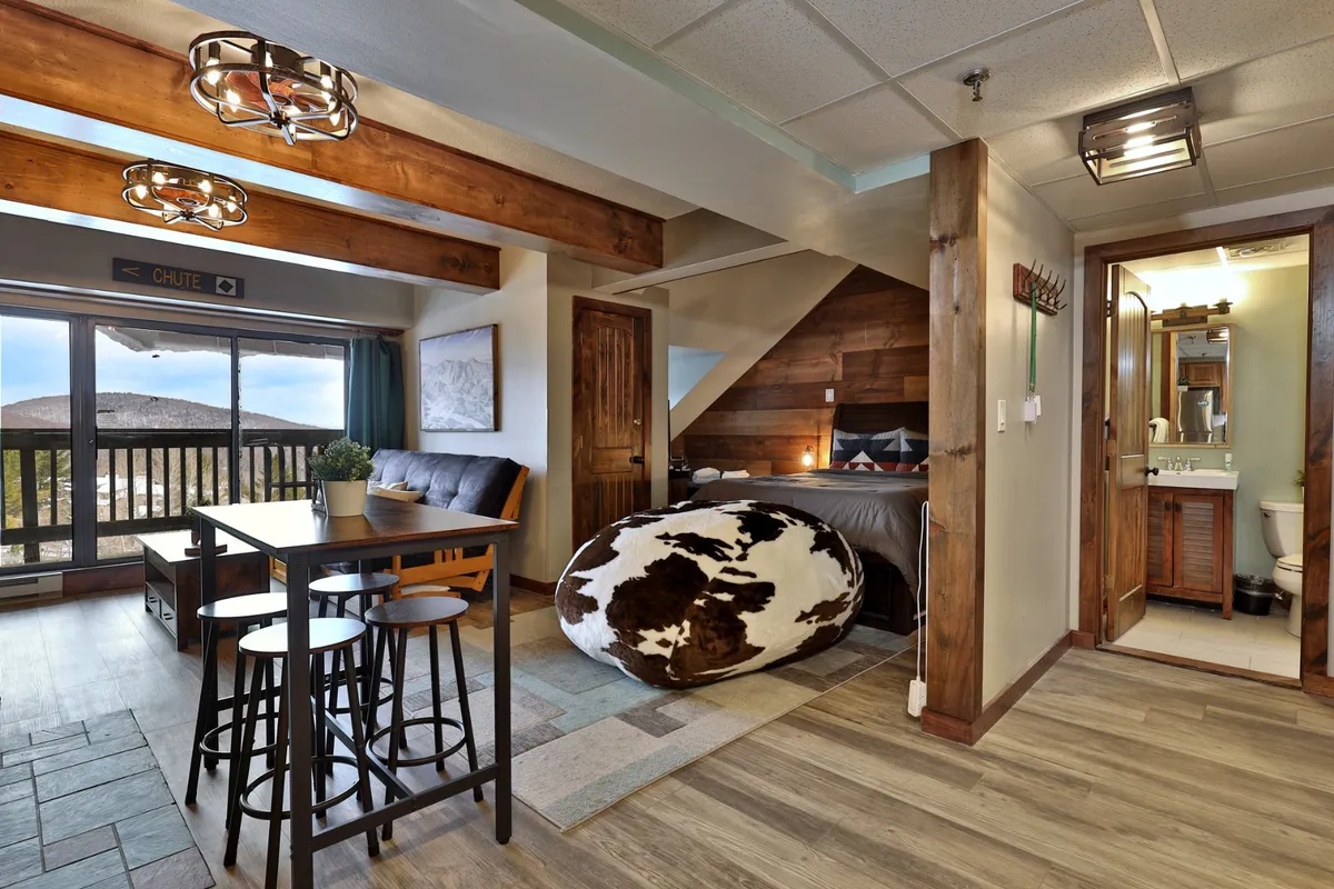 A bright, inviting space with a dining table, cowhide bean bag, rustic bed, and large windows offering views of the outdoors. The blend of comfortable furnishings and natural light highlights the connection between indoor relaxation and the accessible natural surroundings.
