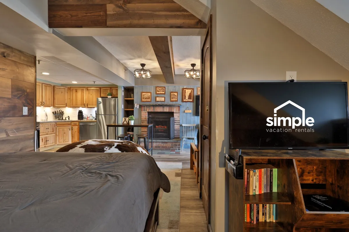 A spacious, open studio layout with a comfortable bed, a fireplace emitting a soft glow, a wall-mounted TV, rustic wooden beams, and a compact kitchen area in view. The space is designed for both unwinding and convenience.