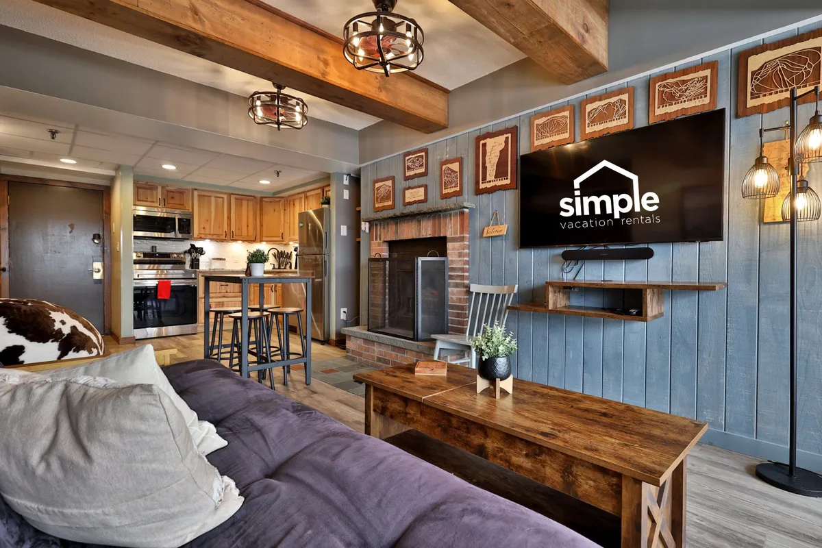This inviting interior features a plush sofa set in front of a glowing electric fireplace, with rustic wooden beams overhead and a Smart TV prominently displayed. A dining area is set to the side, and decorative wall art adds a touch of mountain charm.