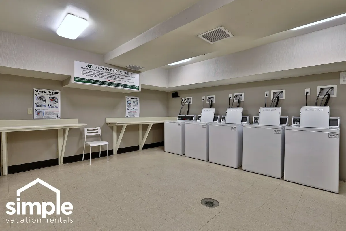 A practical shared laundry room featuring modern washing machines, a drying area, information posters, and a simple white chair. The tidy, well-lit space makes daily routines easy and convenient during a resort stay.