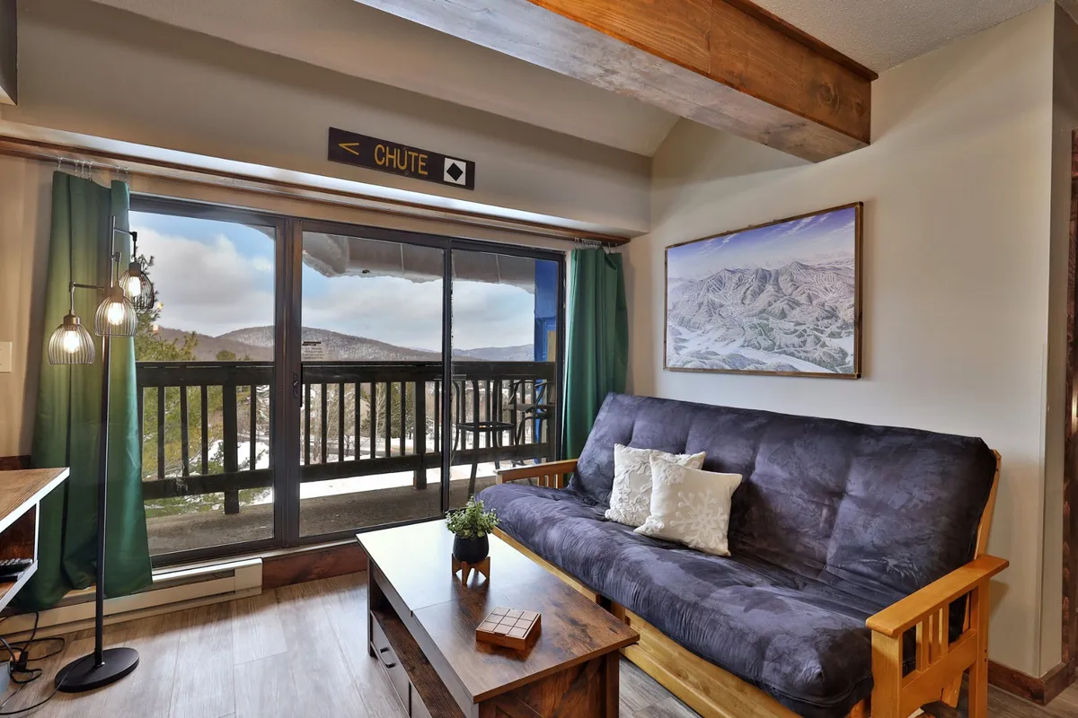 A living area with a futon sofa facing a large window framing scenic mountain views. The space is cozy and comfortable, enhanced by wooden furniture and mountain-themed artwork, blending indoor comfort with the allure of the outdoors.