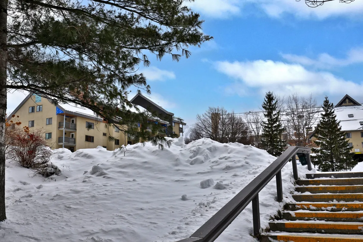 This image showcases the exterior of the property with a snow-covered ground, staircase, surrounding trees, and nearby buildings, evoking the feeling of arriving at a winter destination. The scene hints at a vibrant après-ski environment just steps away from the serenity of the accommodation.