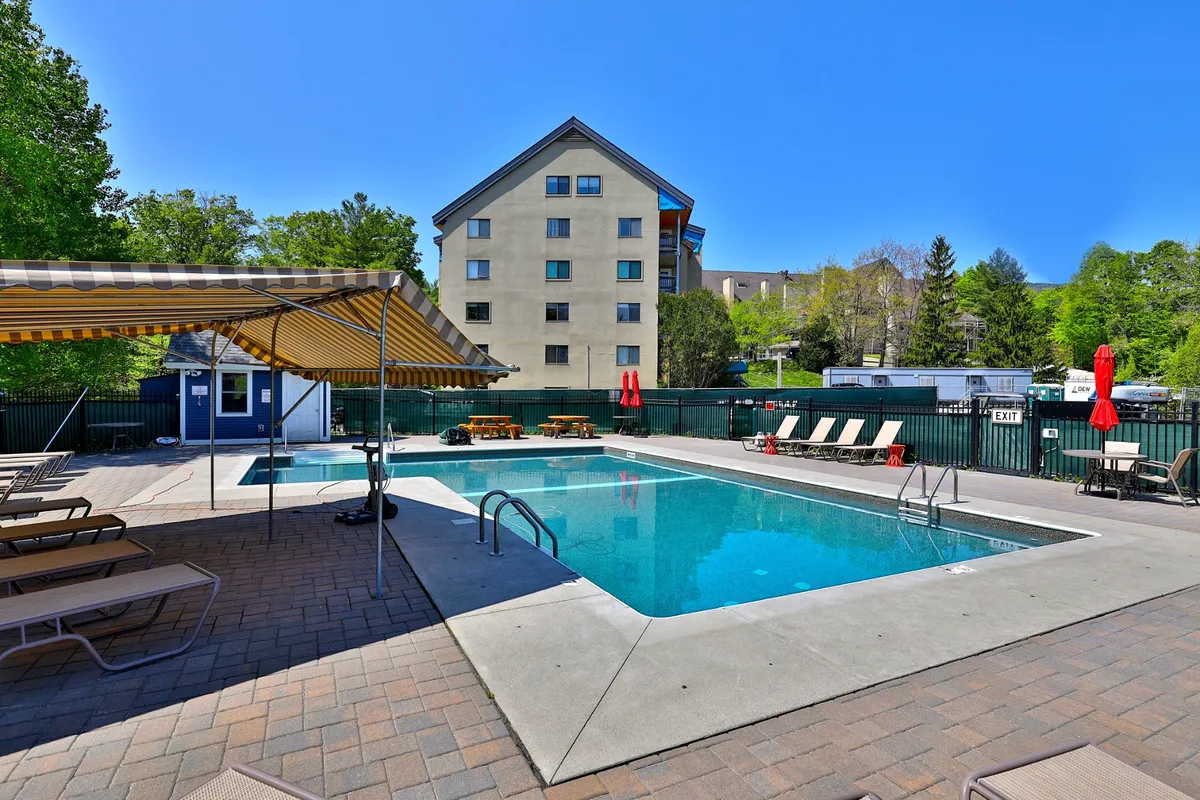 A sunlit outdoor swimming pool surrounded by lounge chairs, with an awning and the building visible in the background. The pool area is inviting and clearly designed for relaxation and social gatherings in the warmer months.