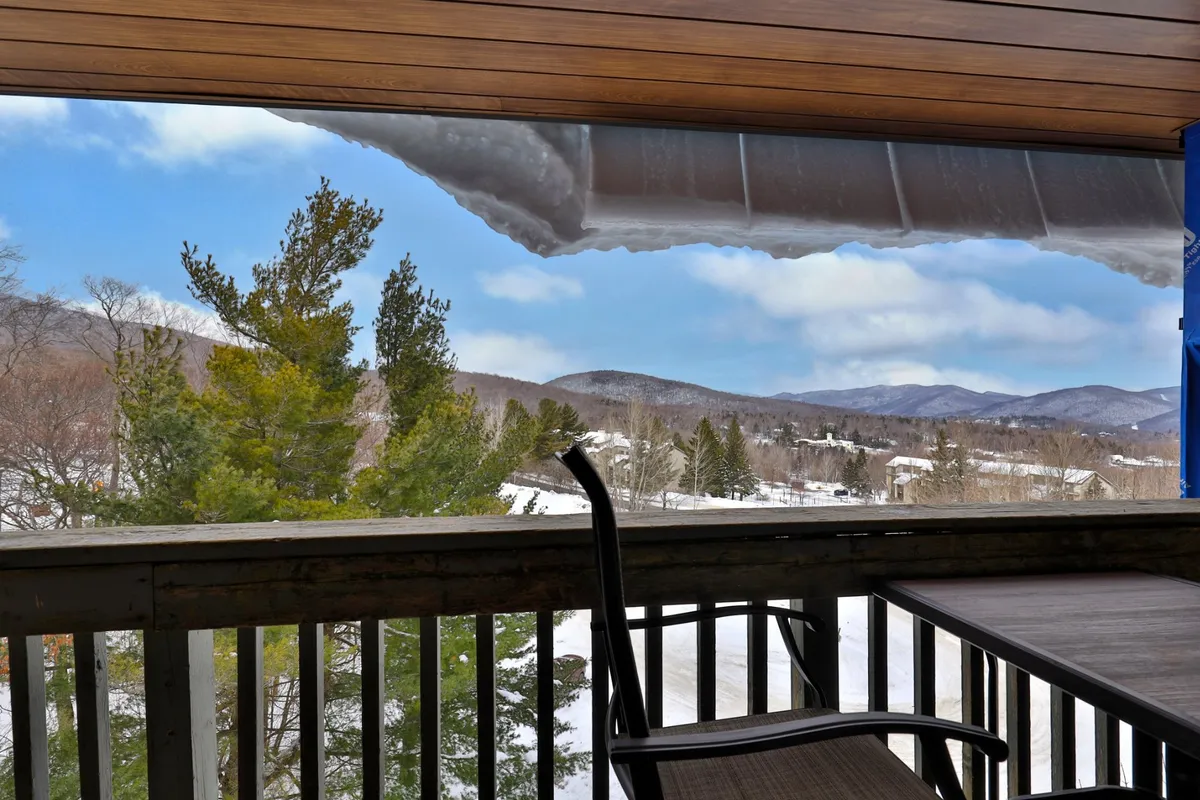 This image captures a tranquil winter scene with snow-covered mountains, a frozen landscape, green pine trees, and a wooden balcony railing. The natural beauty is visible from an outdoor vantage point, inviting guests to step out and take in the fresh mountain air.