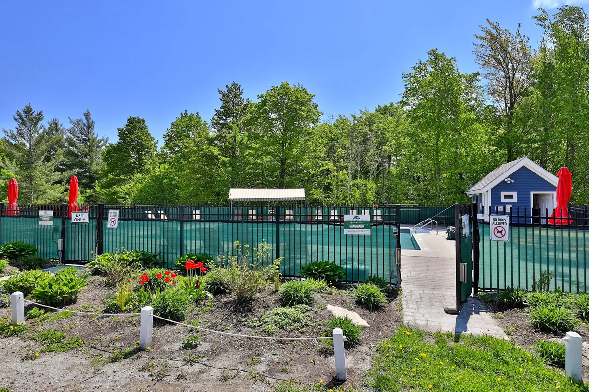 A vibrant exterior pool scene featuring a swimming pool with red umbrellas, landscaped gardens, and a fenced area. The space is lively and offers both relaxation and opportunities for socializing or community events.